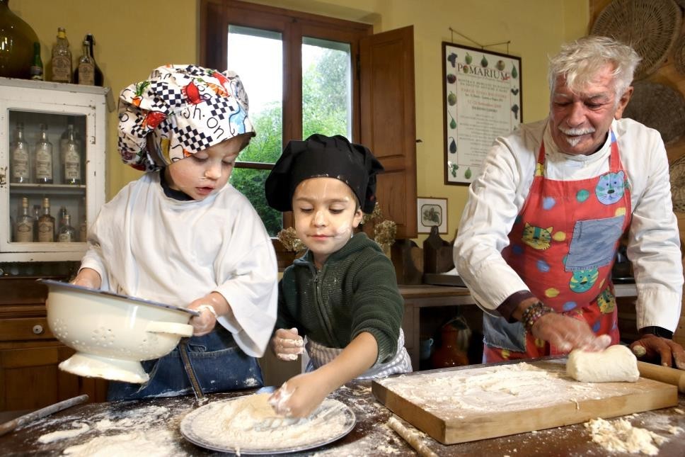 Bambini con le mani in pasta osservati da un nonno intento anche lui a preparare cibo.