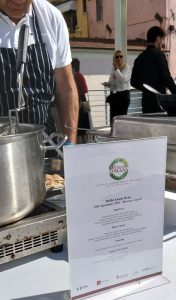 Un cuoco dietro una pentola e in primo piano il menu con il logo di Vetrina Toscana